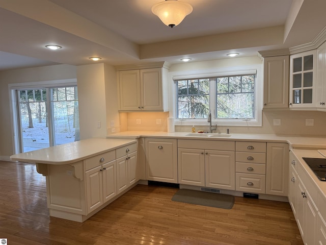 kitchen featuring wood finished floors, a peninsula, a sink, light countertops, and glass insert cabinets