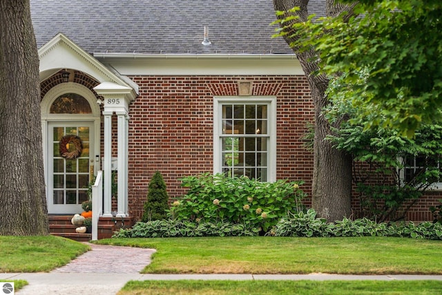 view of exterior entry with brick siding, a lawn, and a shingled roof