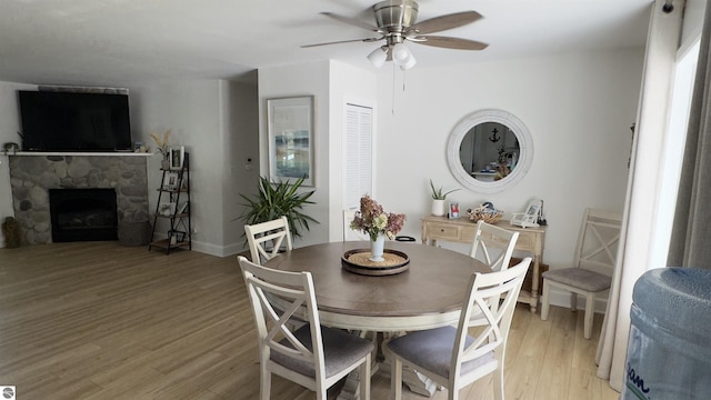 dining area featuring light wood finished floors, a stone fireplace, baseboards, and a ceiling fan