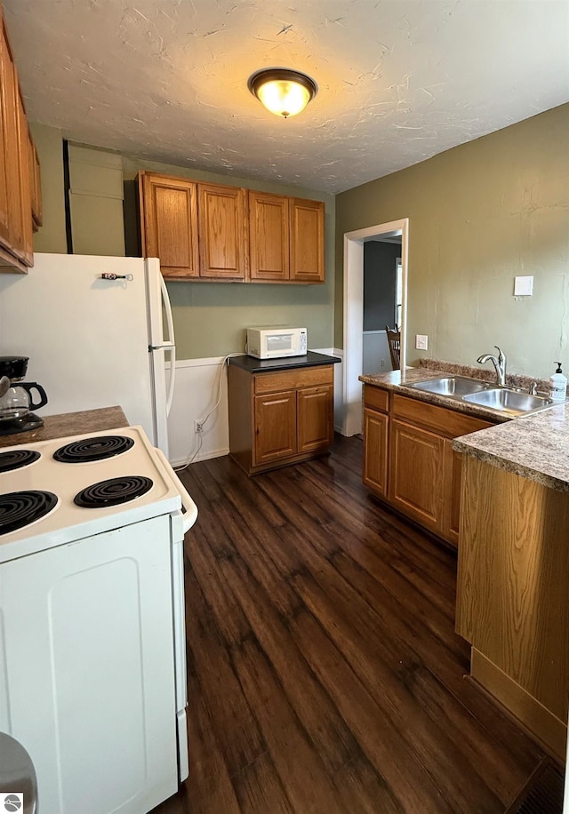 kitchen with a sink, a textured ceiling, dark wood-style floors, white appliances, and brown cabinetry