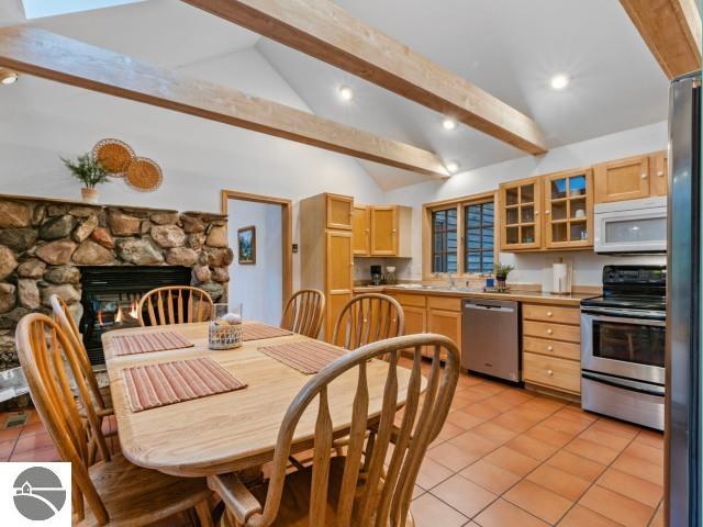 kitchen featuring light brown cabinetry, lofted ceiling with beams, stainless steel appliances, light tile patterned flooring, and glass insert cabinets