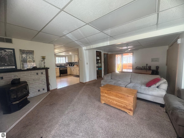 living area featuring a wood stove, visible vents, a ceiling fan, light carpet, and a paneled ceiling