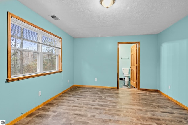 unfurnished bedroom featuring a textured ceiling, ensuite bath, visible vents, wood finished floors, and baseboards