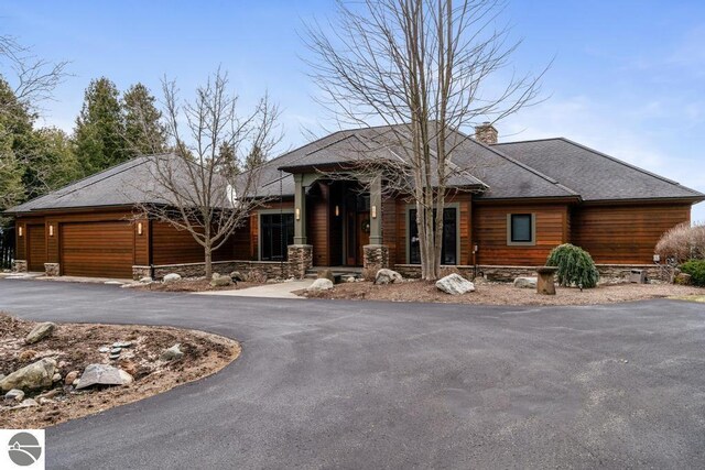 6579 Woods Creek Dr, Charlevoix, MI 49720 - prairie-style house featuring roof with shingles, stone siding, an attached garage, a chimney, and driveway