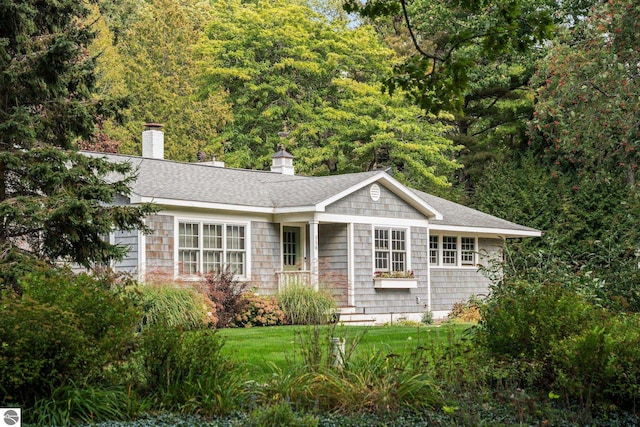 ranch-style house with roof with shingles, a chimney, a forest view, and a front yard