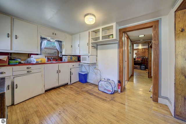 kitchen with white microwave, white cabinetry, open shelves, light wood finished floors, and a sink