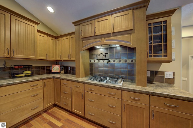 kitchen featuring glass insert cabinets, light wood-style floors, stainless steel gas stovetop, vaulted ceiling, and decorative backsplash