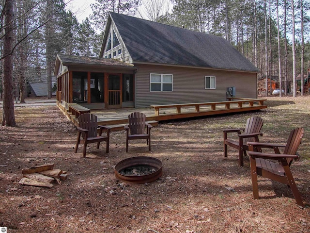 rear view of house featuring a wooden deck, a sunroom, a shingled roof, and an outdoor fire pit