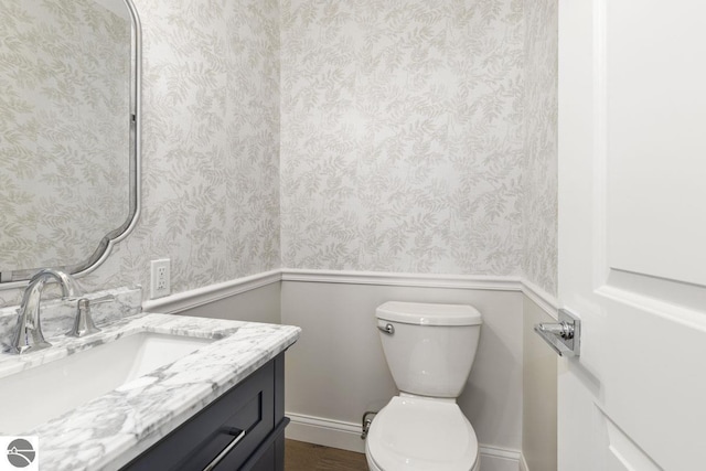 half bath with wallpapered walls, a wainscoted wall, vanity, and toilet