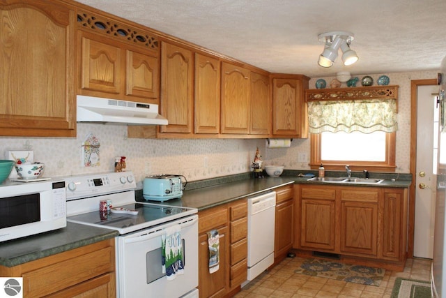 kitchen featuring brown cabinetry, dark countertops, a sink, under cabinet range hood, and white appliances