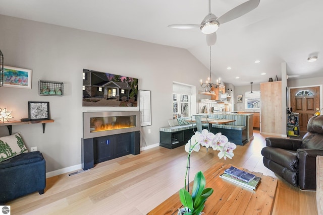 living area with a glass covered fireplace, ceiling fan with notable chandelier, baseboards, lofted ceiling, and light wood-type flooring