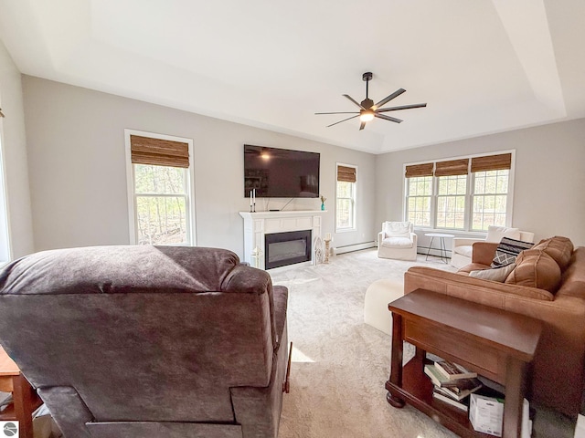 living room with light carpet, a glass covered fireplace, a ceiling fan, a baseboard radiator, and a tray ceiling