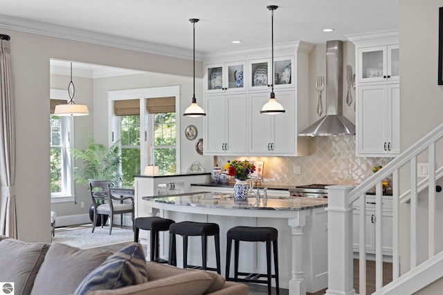 kitchen featuring wall chimney range hood, white cabinets, a kitchen breakfast bar, dark stone counters, and decorative backsplash