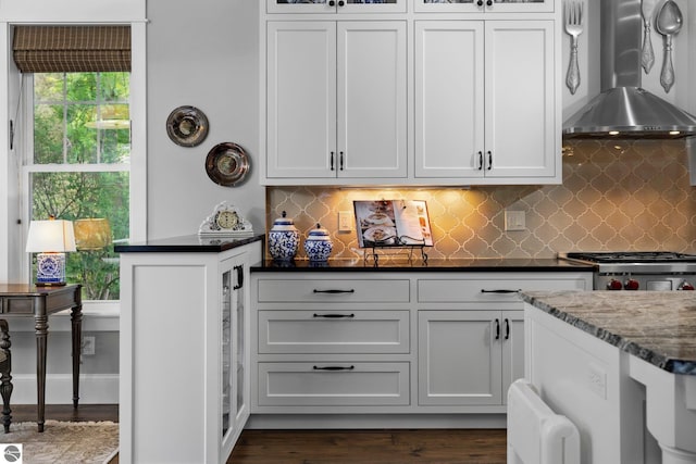kitchen with dark wood finished floors, stove, wall chimney exhaust hood, backsplash, and glass insert cabinets