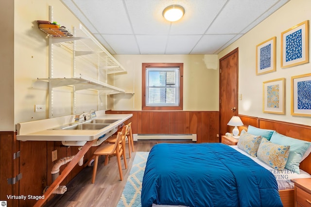 bedroom with a wainscoted wall, a baseboard radiator, a drop ceiling, and wood finished floors