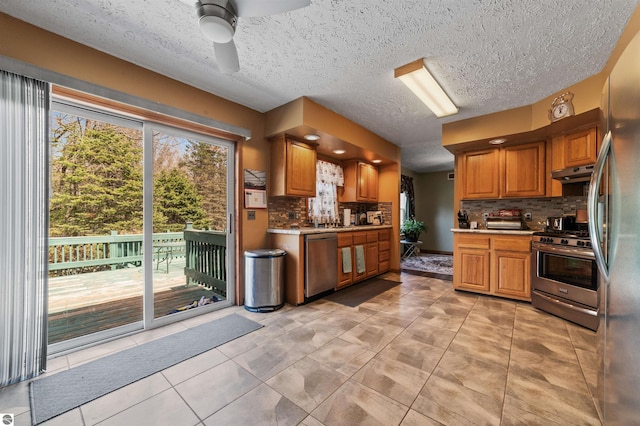 kitchen with decorative backsplash, brown cabinetry, a ceiling fan, under cabinet range hood, and stainless steel appliances
