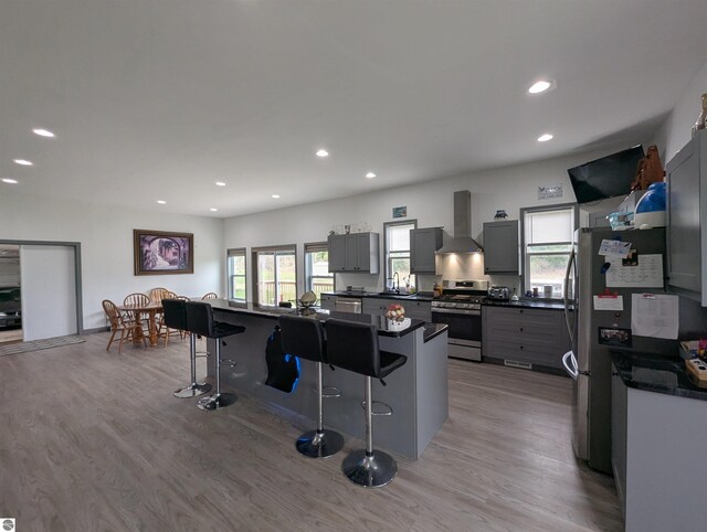 kitchen featuring wall chimney range hood, dark countertops, appliances with stainless steel finishes, a kitchen breakfast bar, and light wood-type flooring