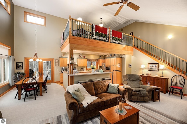 living area featuring stairs, baseboards, high vaulted ceiling, light colored carpet, and ceiling fan with notable chandelier