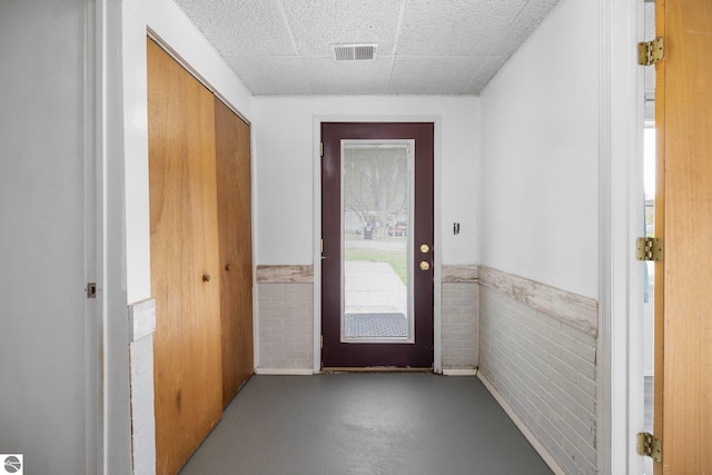 doorway featuring concrete flooring, a wainscoted wall, and a drop ceiling