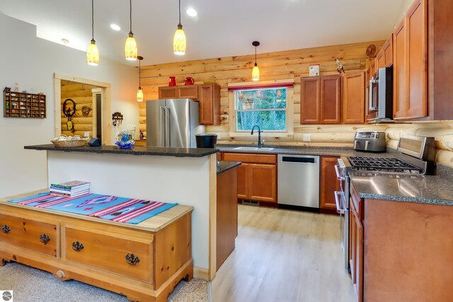 kitchen with appliances with stainless steel finishes, a sink, light wood-type flooring, hanging light fixtures, and dark stone countertops