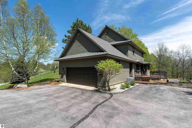 view of side of property featuring driveway, a wooden deck, a garage, and a shingled roof