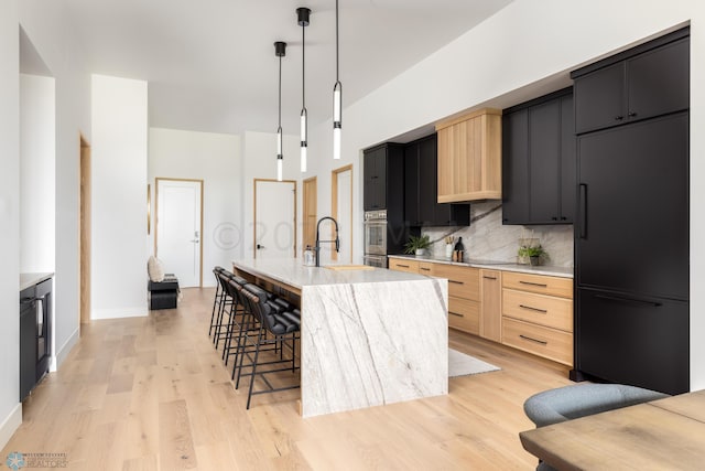 kitchen featuring light hardwood / wood-style floors, backsplash, decorative light fixtures, black refrigerator, and an island with sink