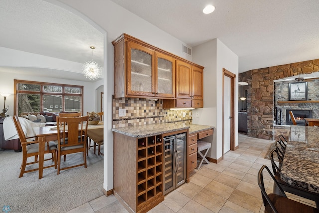 kitchen featuring wine cooler, light stone counters, light colored carpet, decorative backsplash, and a stone fireplace