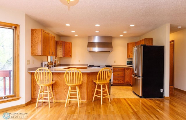 kitchen featuring wall chimney range hood, a textured ceiling, light hardwood / wood-style flooring, stainless steel appliances, and a kitchen bar