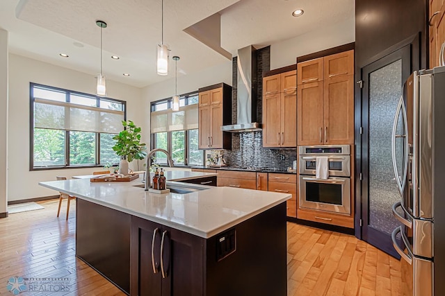 kitchen with light hardwood / wood-style floors, an island with sink, decorative light fixtures, wall chimney range hood, and appliances with stainless steel finishes