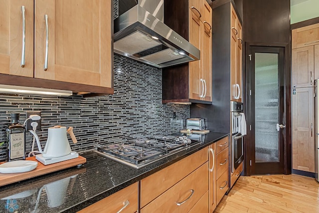 kitchen with appliances with stainless steel finishes, wall chimney exhaust hood, tasteful backsplash, and light wood-type flooring