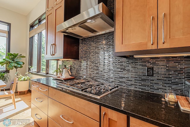 kitchen featuring dark stone counters, wall chimney exhaust hood, stainless steel gas cooktop, and tasteful backsplash