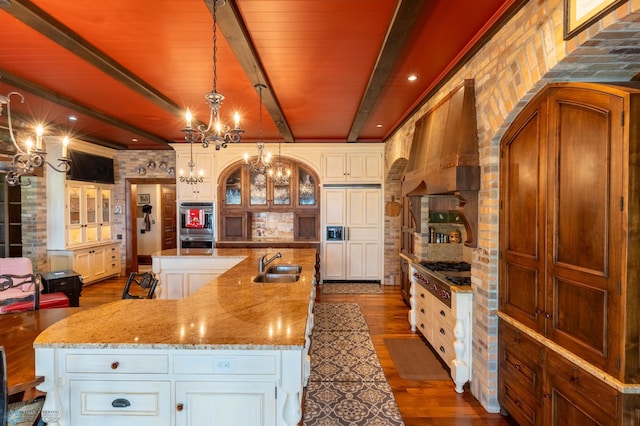 kitchen featuring beamed ceiling, a center island with sink, wall chimney range hood, sink, and white cabinets