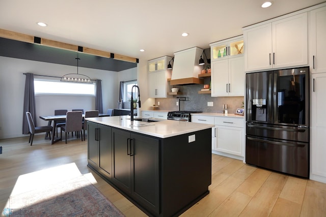 kitchen with white cabinets, premium range hood, black fridge with ice dispenser, decorative backsplash, and a center island with sink