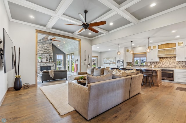 living room featuring beamed ceiling, light hardwood / wood-style flooring, a stone fireplace, ceiling fan, and coffered ceiling