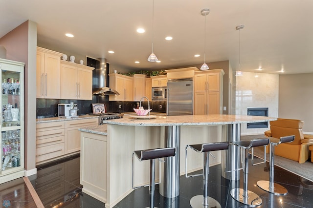 kitchen with hanging light fixtures, wall chimney range hood, a center island with sink, and built in appliances