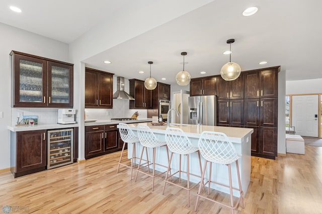 kitchen featuring wall chimney range hood, wine cooler, appliances with stainless steel finishes, light hardwood / wood-style flooring, and pendant lighting