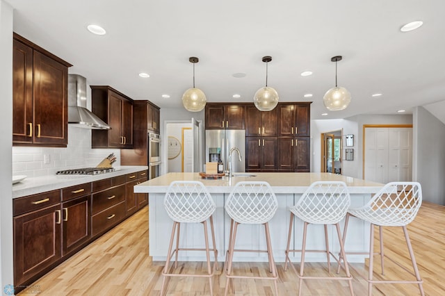 kitchen featuring wall chimney exhaust hood, appliances with stainless steel finishes, a kitchen island with sink, and hanging light fixtures