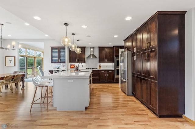 kitchen with wall chimney range hood, appliances with stainless steel finishes, light hardwood / wood-style floors, pendant lighting, and a center island with sink