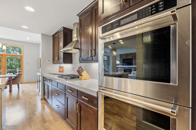 kitchen with stainless steel appliances, dark brown cabinetry, wall chimney exhaust hood, decorative backsplash, and light hardwood / wood-style flooring