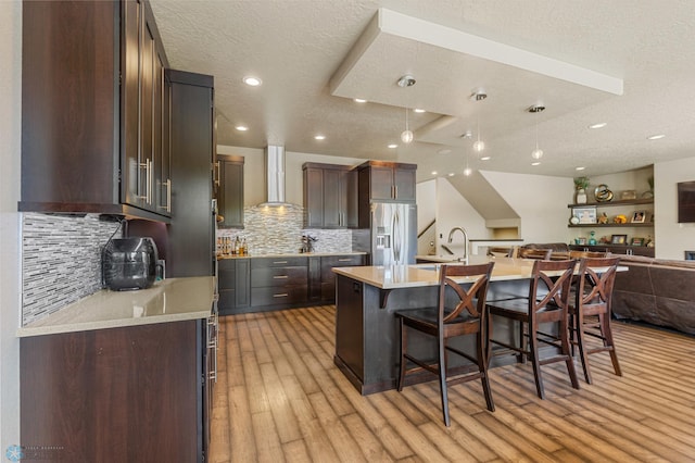 kitchen featuring a textured ceiling, a center island with sink, light hardwood / wood-style flooring, stainless steel appliances, and wall chimney range hood