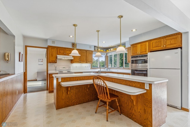 kitchen with hanging light fixtures, white appliances, sink, a breakfast bar area, and a kitchen island with sink