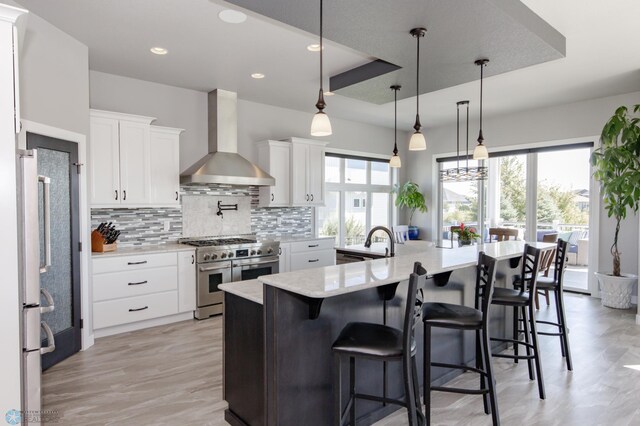 kitchen featuring high end stove, wall chimney range hood, a raised ceiling, and white cabinets