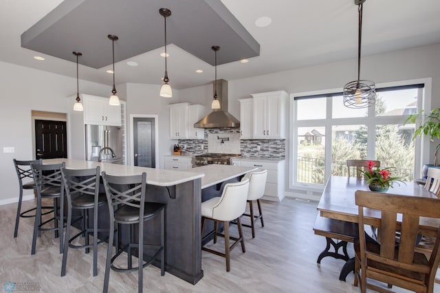 kitchen with pendant lighting, a large island with sink, wall chimney range hood, and white cabinetry