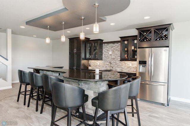 bar with dark stone countertops, stainless steel refrigerator with ice dispenser, hanging light fixtures, dark brown cabinets, and backsplash