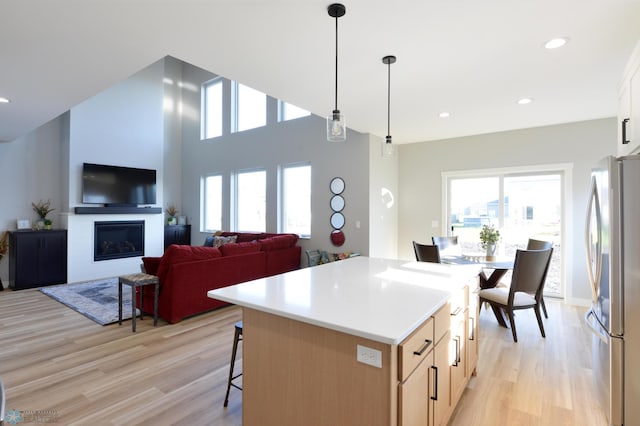 kitchen with hanging light fixtures, stainless steel refrigerator, a kitchen island, light wood-type flooring, and light brown cabinetry