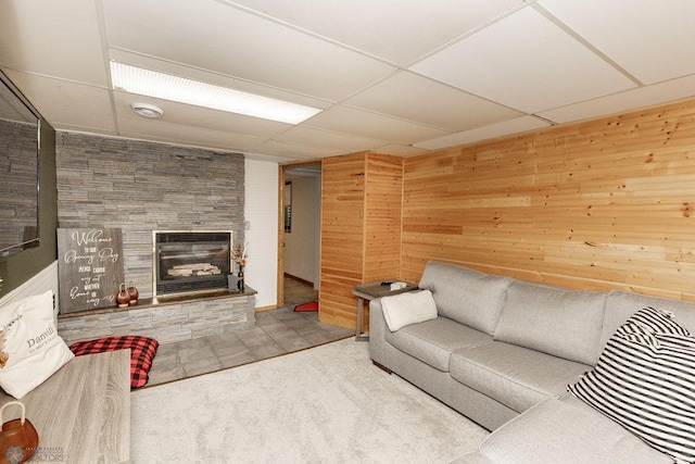 carpeted living room with a stone fireplace, a drop ceiling, and wood walls