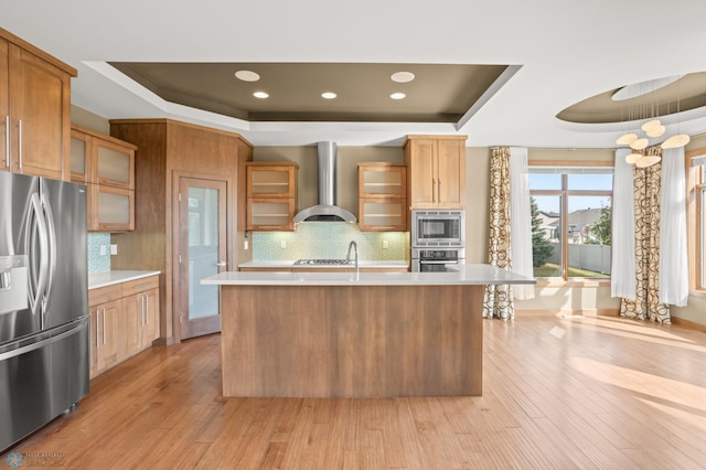 kitchen with a center island with sink, appliances with stainless steel finishes, a raised ceiling, and wall chimney range hood