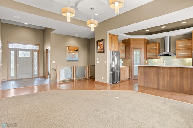 kitchen with hanging light fixtures, backsplash, wall chimney exhaust hood, light wood-type flooring, and stainless steel refrigerator with ice dispenser