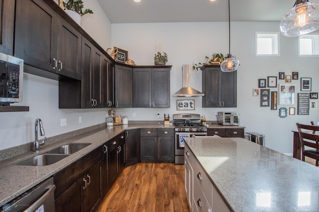 kitchen featuring sink, hanging light fixtures, wall chimney range hood, appliances with stainless steel finishes, and light stone countertops