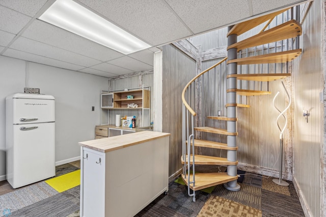 interior space with a drop ceiling, wood walls, dark colored carpet, and white fridge